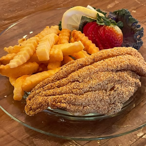 Fried catfish and crinkle-cut fries with a lemon wedge, strawberry, and kale on a glass plate.